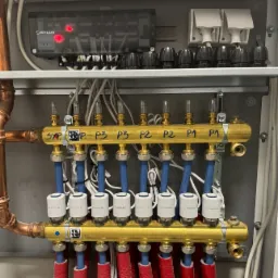 Rozdzielacz ogrzewania podłogowego w szafce, z podłączonymi rurami w izolacji czerwonej, niebieskimi rurami doprowadzającymi i elektronicznym sterownikiem Salus z czerwonymi diodami.