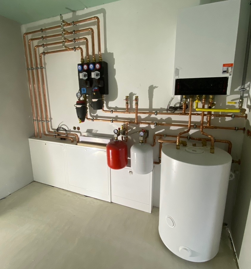 Kompletna instalacja grzewcza z piecem, zasobnikiem i rozdzielaczem, rury miedziane prowadzone po ścianie, widoczne zawory i manometry.