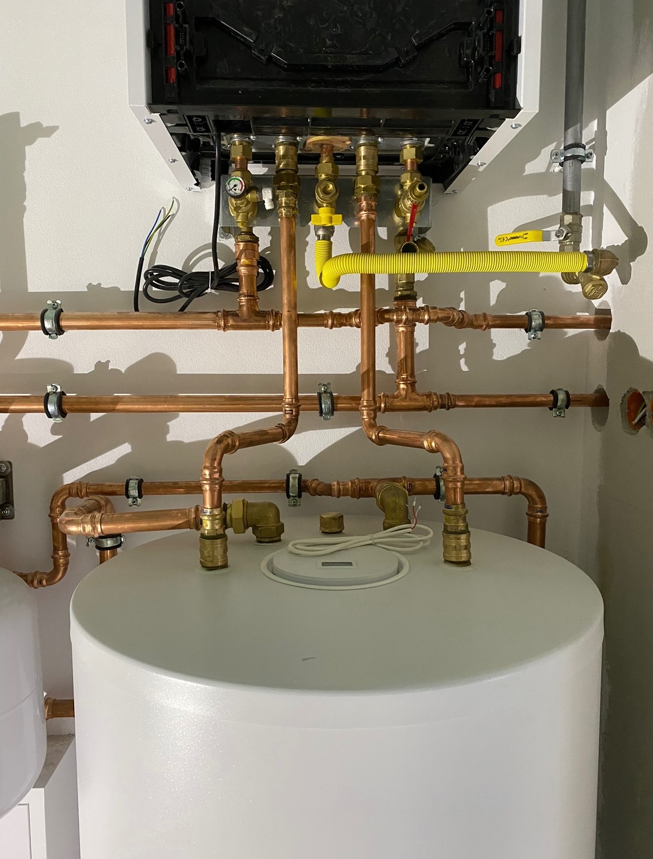 Nowoczesny piec gazowy z podłączeniem miedzianych rur i żółtą elastyczną rurą gazową, zamocowany na białej ścianie, widok z dołu, detale instalacji.