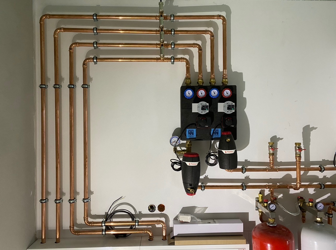 Rozdzielacz ogrzewania podłogowego z miedzianymi rurami i pompami obiegowymi Flamco zamontowany na białej ścianie, widoczne manometry i zawory.