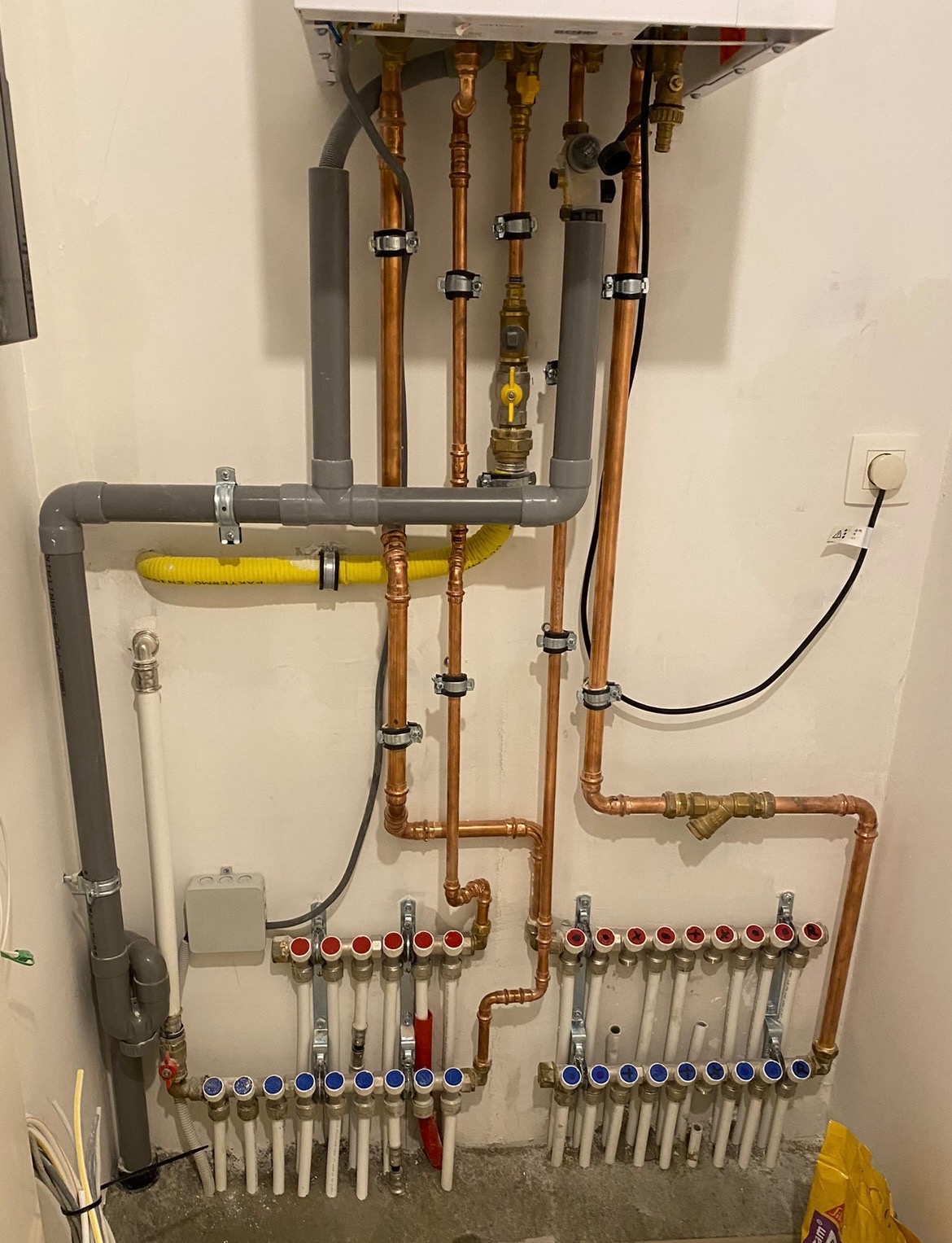 Rozdzielacz ogrzewania podłogowego z piecem gazowym, widoczne miedziane rury i szare rury PCV, żółty przewód gazowy, zamontowane na białej ścianie.