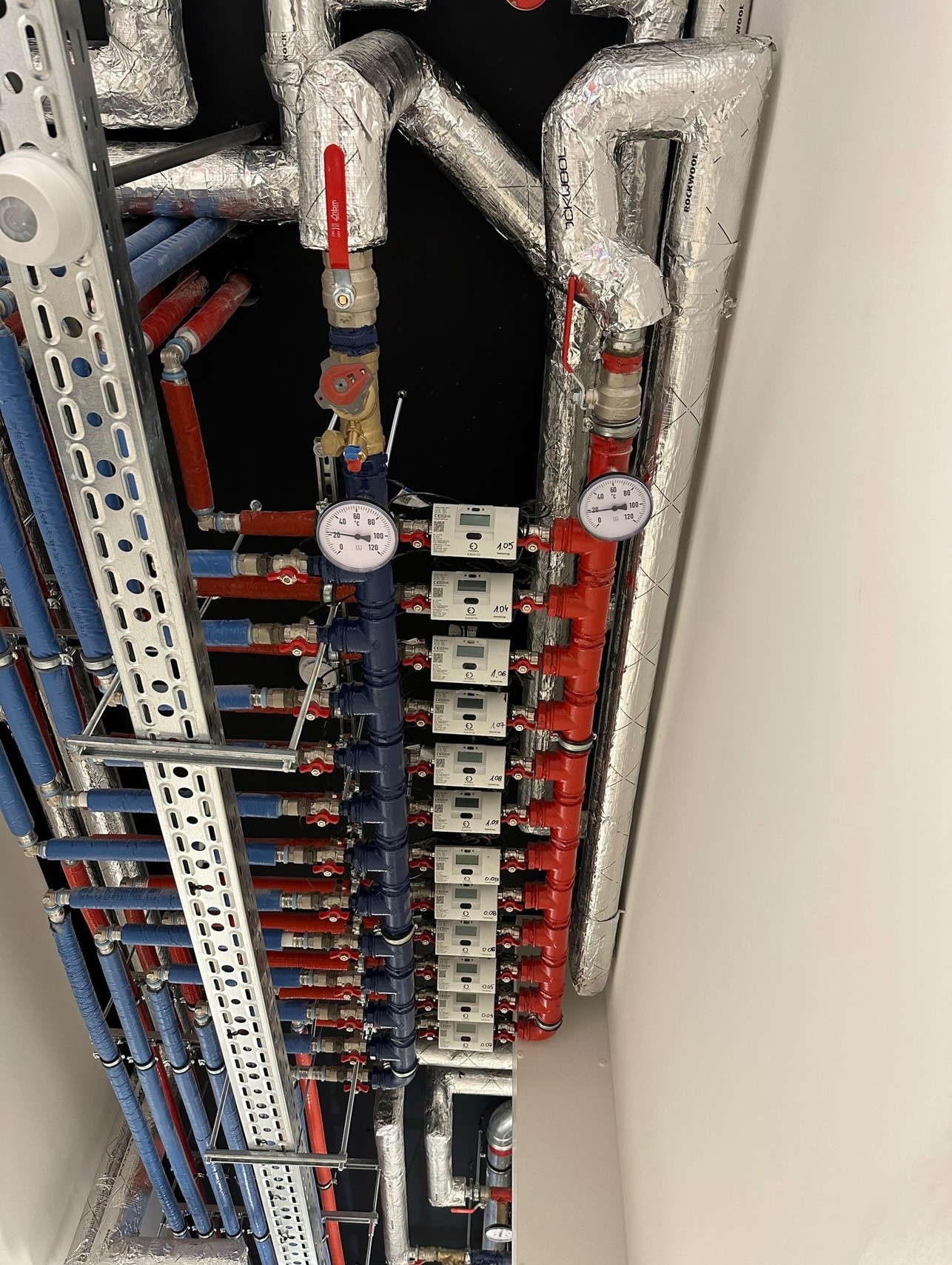 Rozdzielacz instalacji centralnego ogrzewania z licznymi rurami w izolacji, zaworami, licznikami ciepła i manometrami, zamontowany na ścianie w pomieszczeniu technicznym.