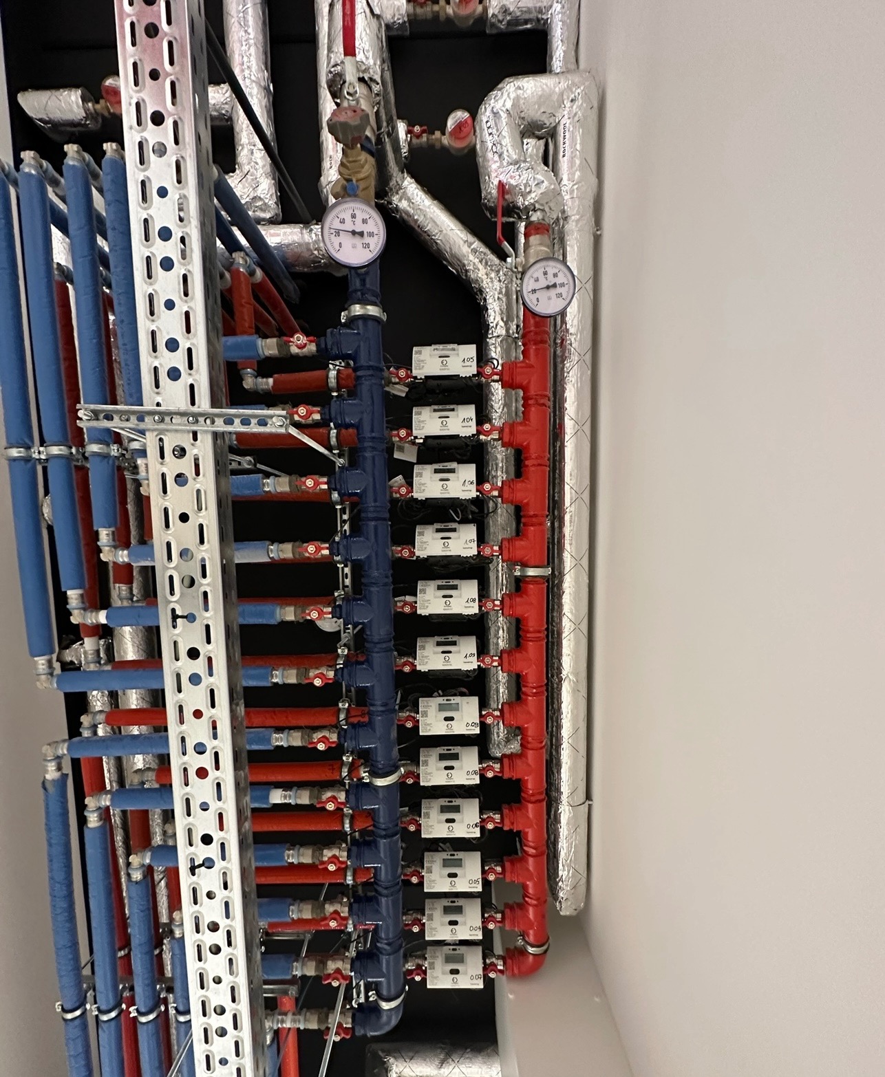 Rozdzielacz instalacji z rurami w kolorach niebieskim i czerwonym, zamontowany na ścianie z widocznymi licznikami ciepła i manometrami, zabezpieczony izolacją.