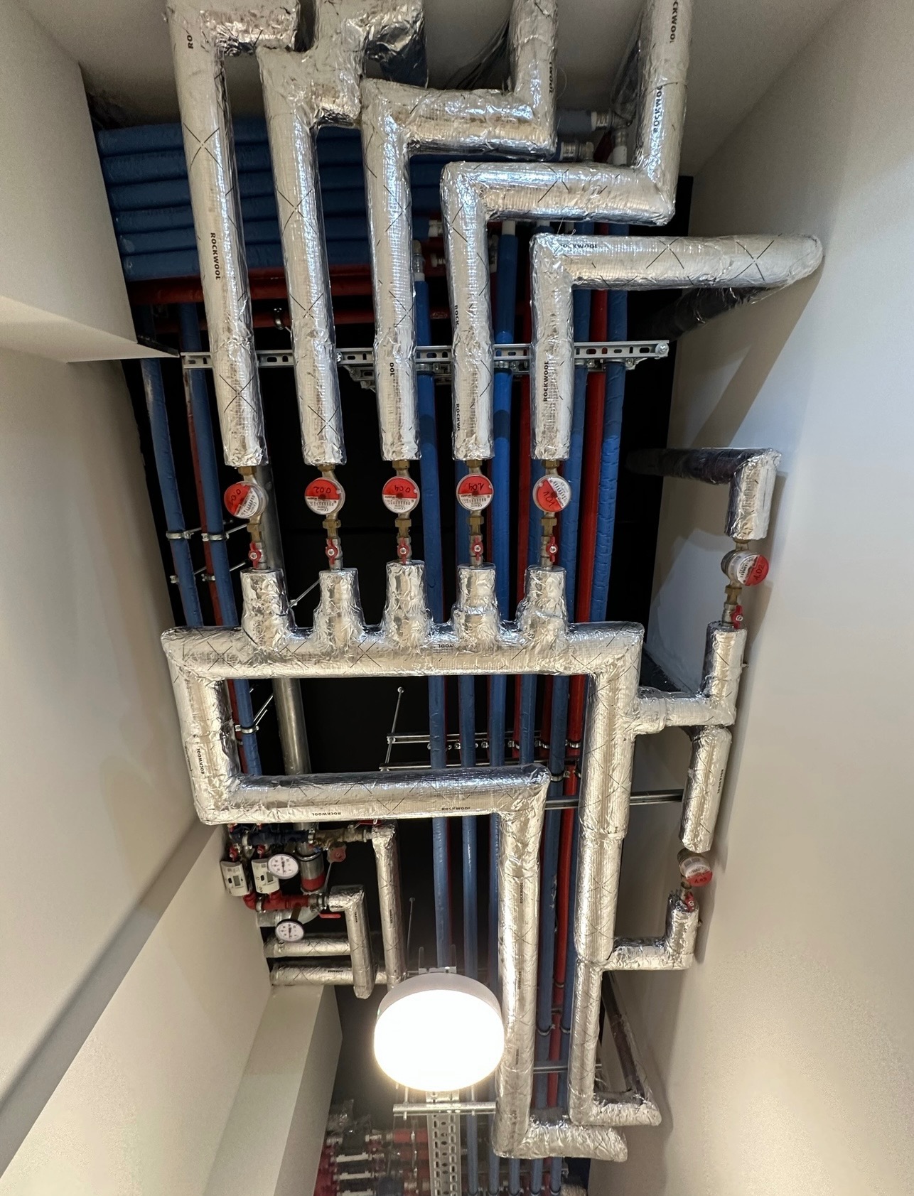 Widok instalacji centralnego ogrzewania i wodnej, z rurami w izolacji aluminiowej i licznikami ciepła, zamontowanymi pod sufitem w nowym budynku.