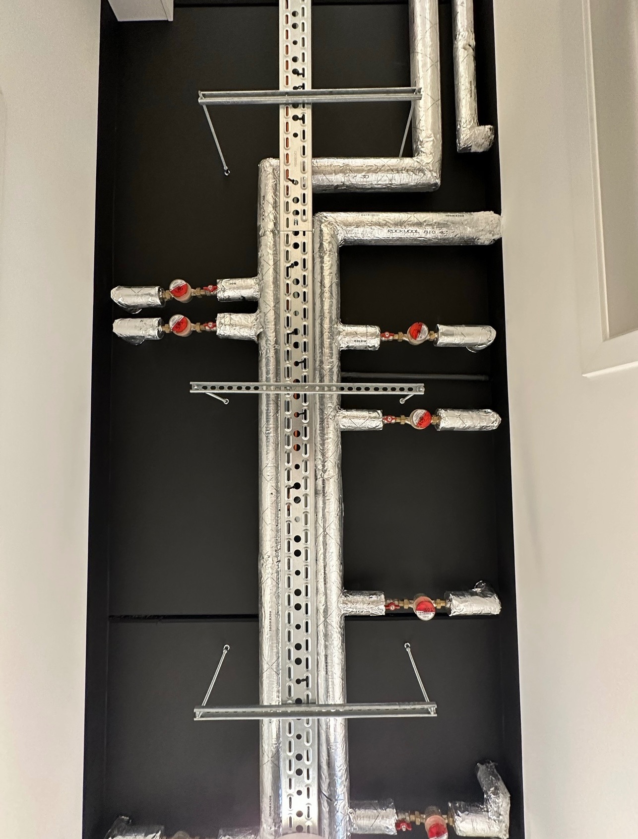 Instalacja rur w izolacji aluminiowej z zamontowanymi zaworami i manometrami, umieszczona w czarnej wnęce ściennej, widok z dołu.