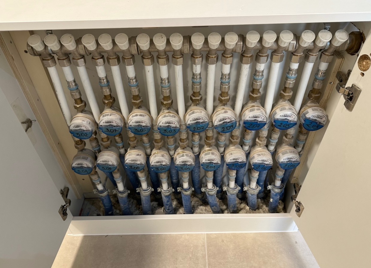 Rozdzielacz ogrzewania podłogowego w szafce, widoczne liczniki z niebieskimi oznaczeniami i numerami pętli grzewczych, białe rury i izolacja.