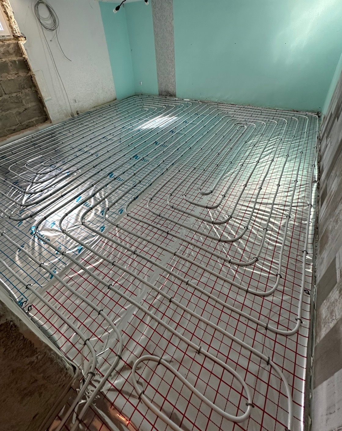 Białe rurki instalacji grzewczej ułożone w pętle na podłodze z folią w kratkę, widoczne fragmenty ścian w trakcie remontu.