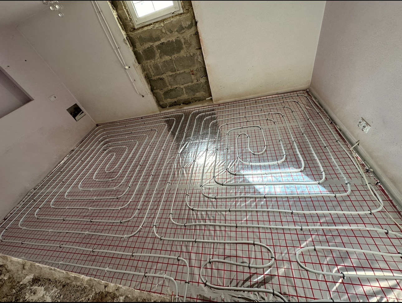 Rozłożone białe rurki na folii z czerwoną siatką, przygotowane do zalania betonem, widok na surowy strop i okno dachowe.