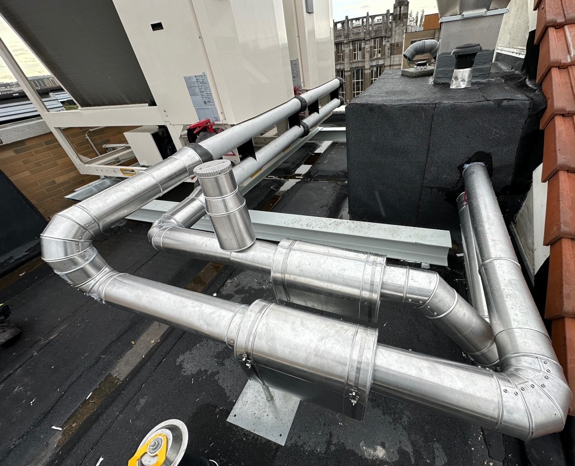 Metalowe rury wentylacyjne na dachu budynku, widoczne elementy systemu wentylacji mechanicznej z odzyskiem ciepła oraz fragment dachu z papy i dachówki.