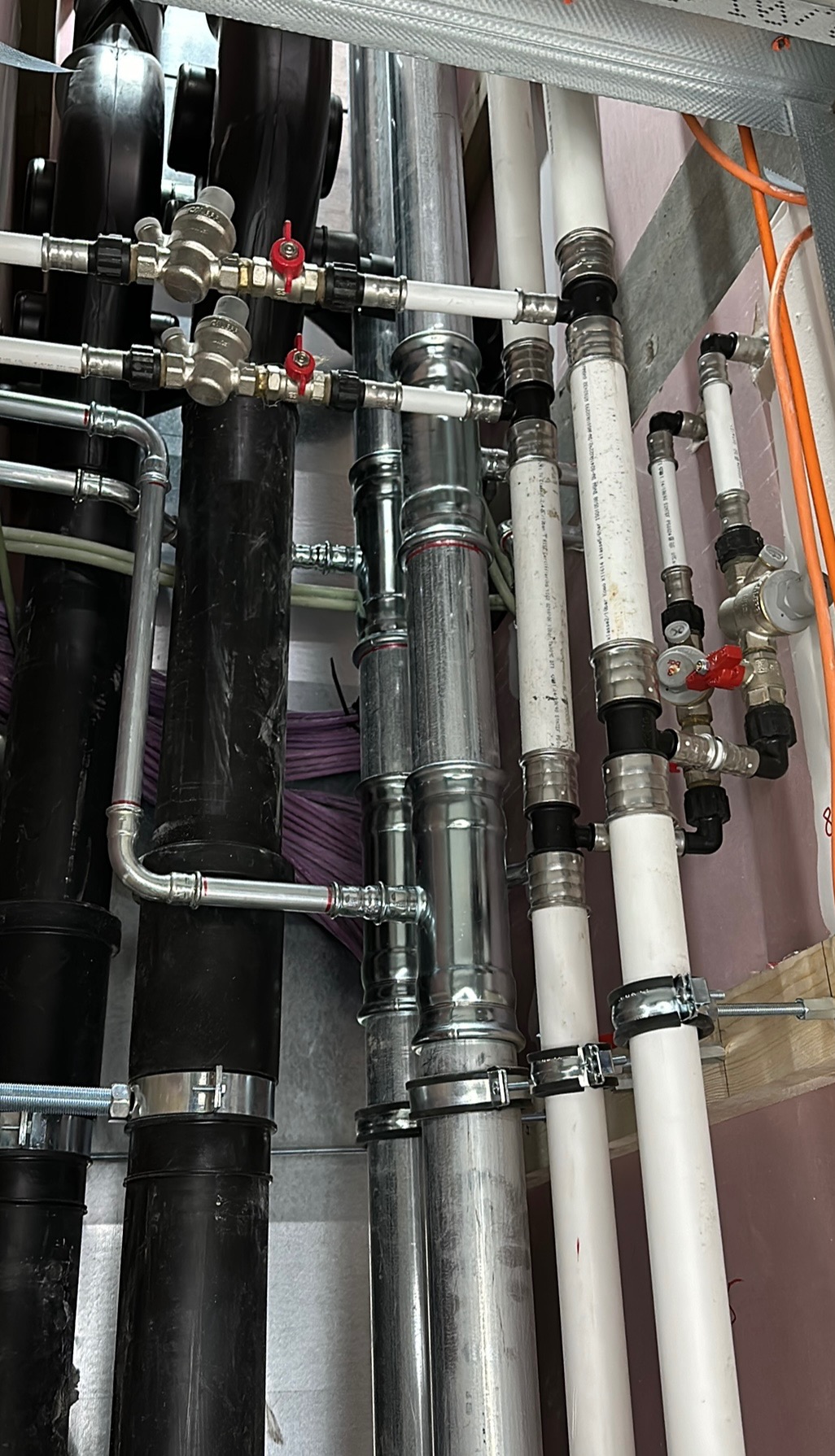 Złożony system rur z zaworami i złączkami, w tym czarne, białe i metalowe rury, przymocowany do ściany z widocznymi przewodami elektrycznymi w tle.