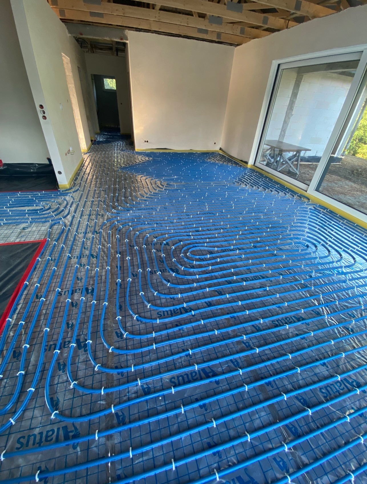Niebieskie rurki ogrzewania podłogowego ułożone na siatce w surowym wnętrzu budynku, widoczne okno i niedokończone ściany.
