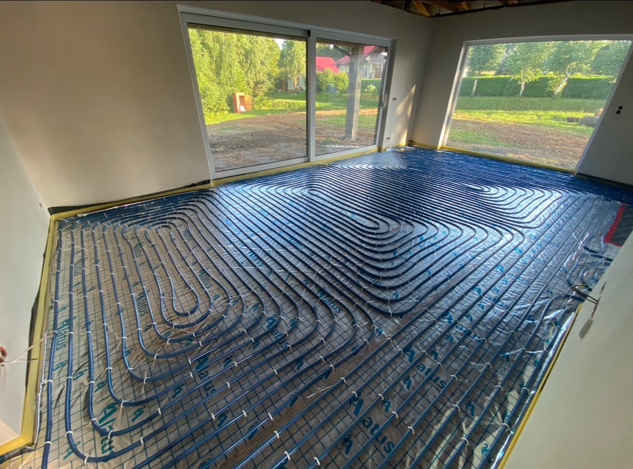 Niebieskie rurki systemu grzewczego podłogowego ułożone spiralnie na folii w nowym domu w Strzyżowie, widoczne okna z widokiem na ogród.