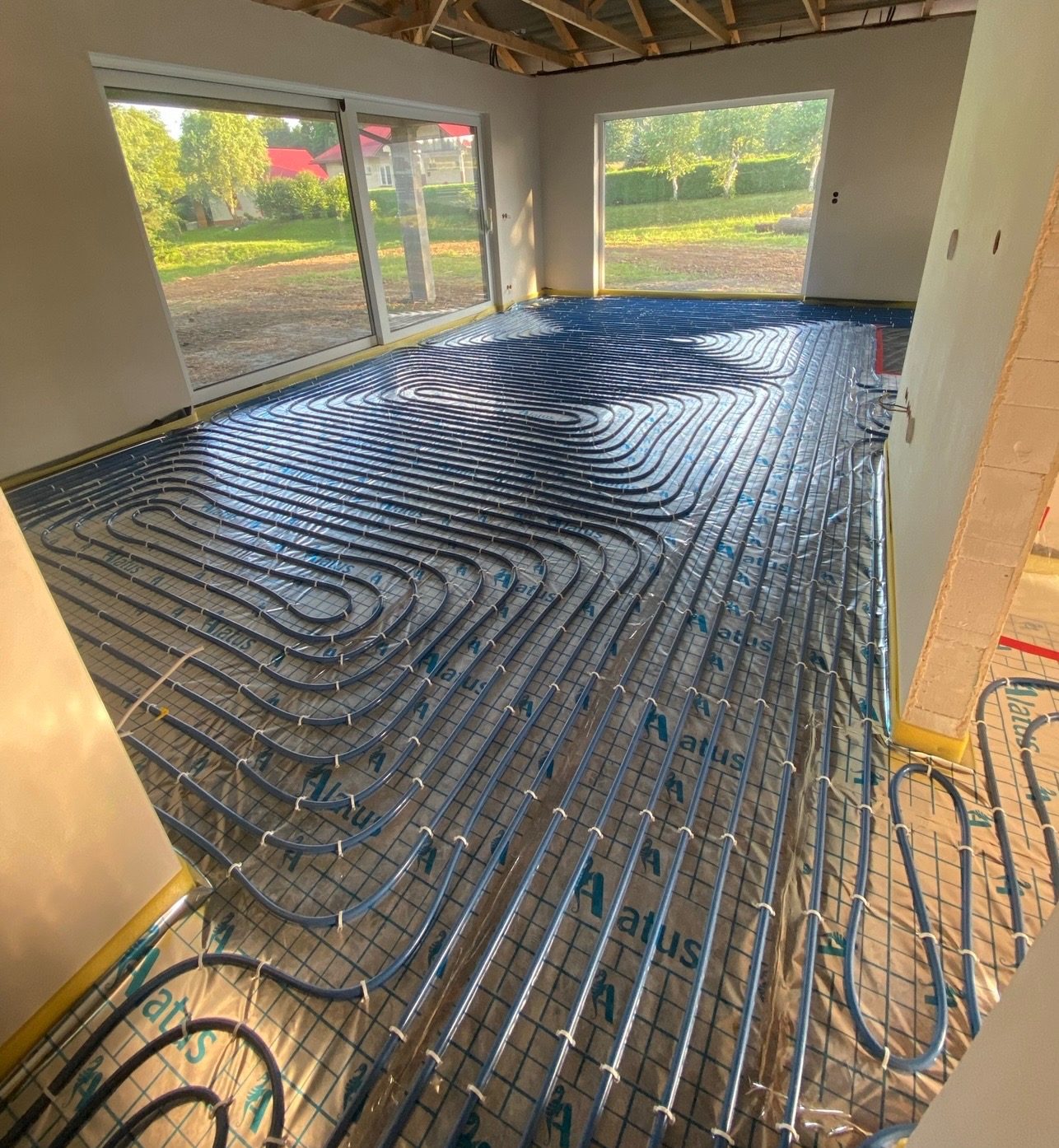 Niebieskie rury ogrzewania podłogowego ułożone na folii z nadrukowanym logo 'Atus' w surowym wnętrzu z dużymi oknami i widokiem na zieleń.