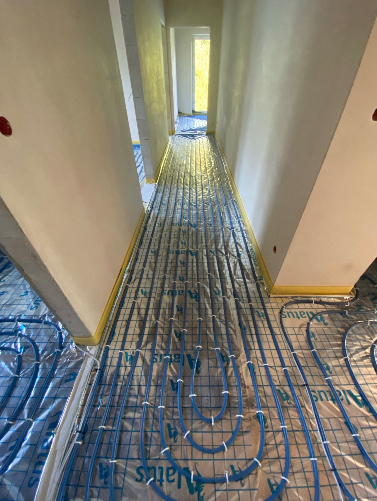 Niebieskie rury grzewcze rozłożone na folii izolacyjnej, przygotowane do zalania wylewką w długim korytarzu z surowymi ścianami, zabezpieczonymi taśmą malarską.