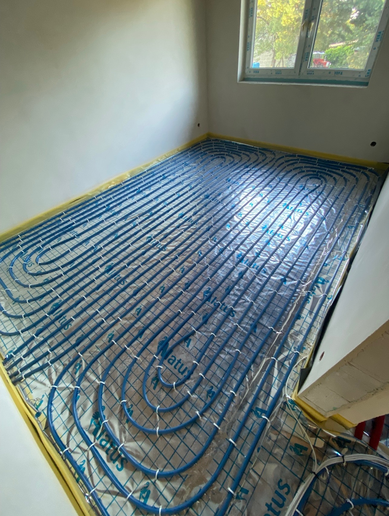 Niebieskie rury układane w pętle na srebrnej folii z nadrukiem w pomieszczeniu przygotowanym pod wylewkę; widoczne okno i żółta taśma zabezpieczająca przy ścianach.