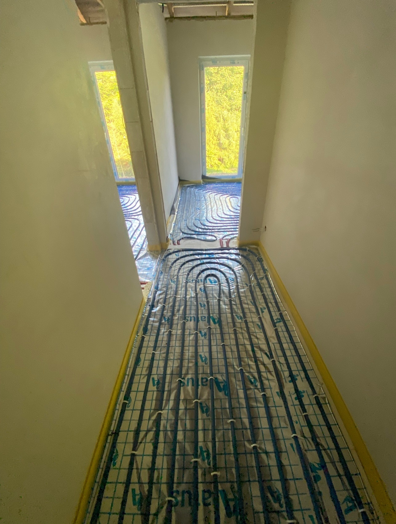 Niedokończona instalacja ogrzewania podłogowego w nowym budynku, rury grzewcze ułożone na folii z siatką, zabezpieczone taśmą malarską przy ścianach, widoczne dwa okna z zielenią na zewnątrz.