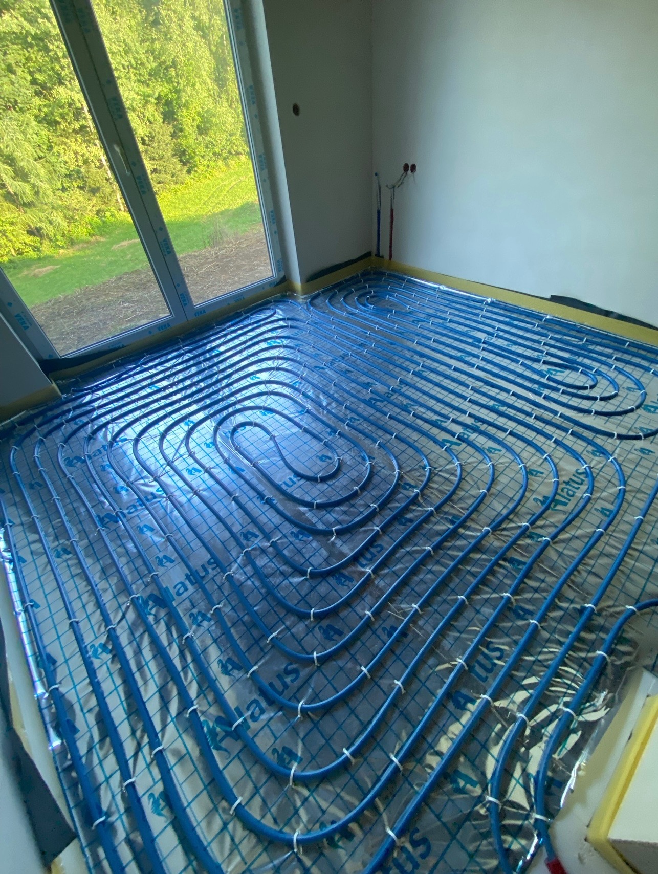 Niebieskie rury instalacji grzewczej podłogowej, ułożone spiralnie na srebrnej folii izolacyjnej w nowym pomieszczeniu z dużym oknem.