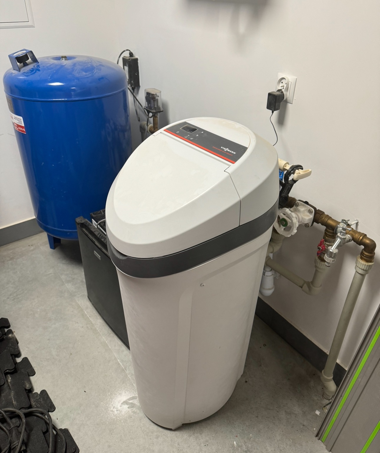 Biały zmiękczacz wody Viessmann z szarym paskiem, podłączony do instalacji hydraulicznej, obok niebieski zbiornik ciśnieniowy, widoczne zasilanie z gniazdka elektrycznego.