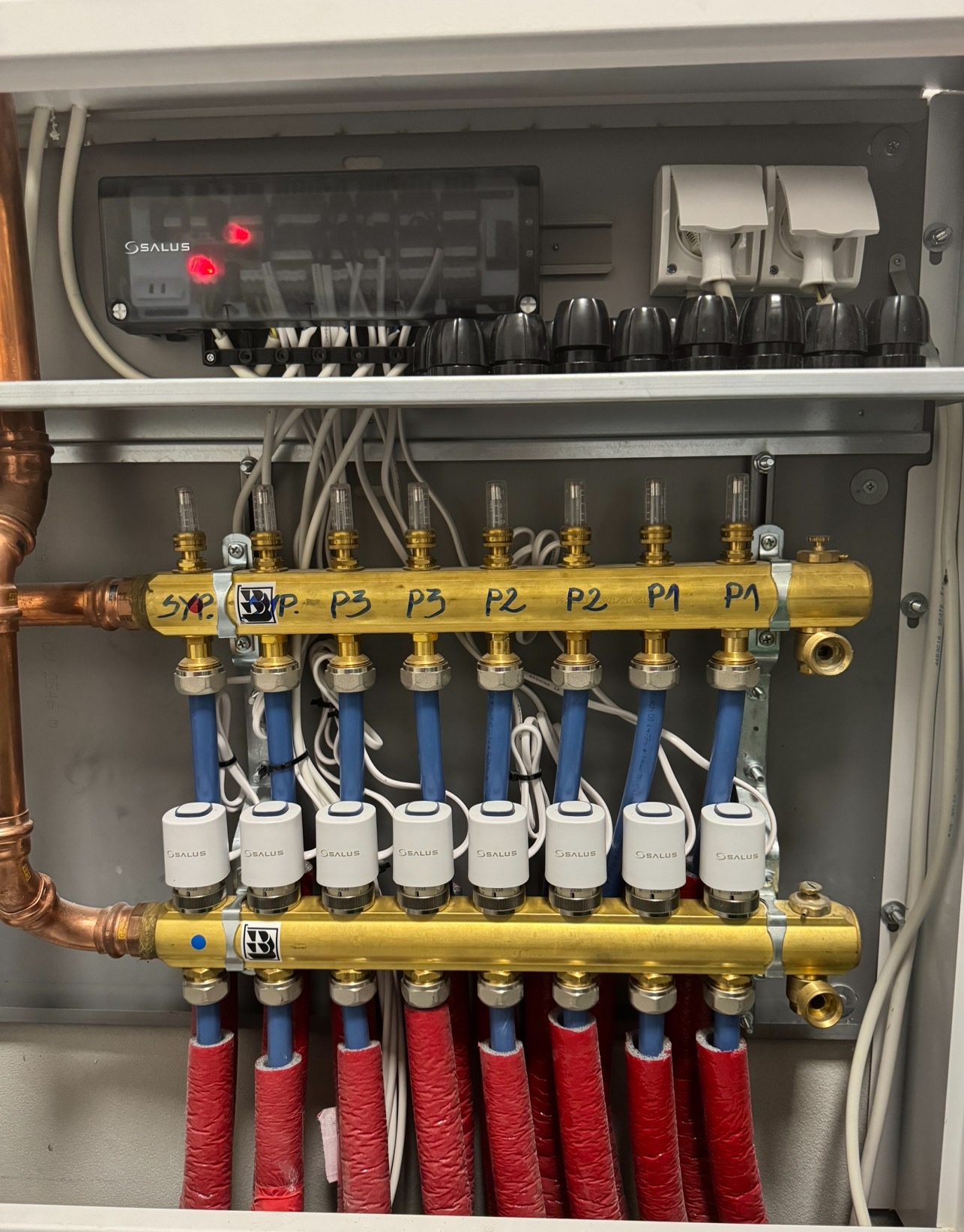 Rozdzielacz ogrzewania podłogowego w szafce, z podłączonymi rurami w izolacji czerwonej, niebieskimi rurami doprowadzającymi i elektronicznym sterownikiem Salus z czerwonymi diodami.