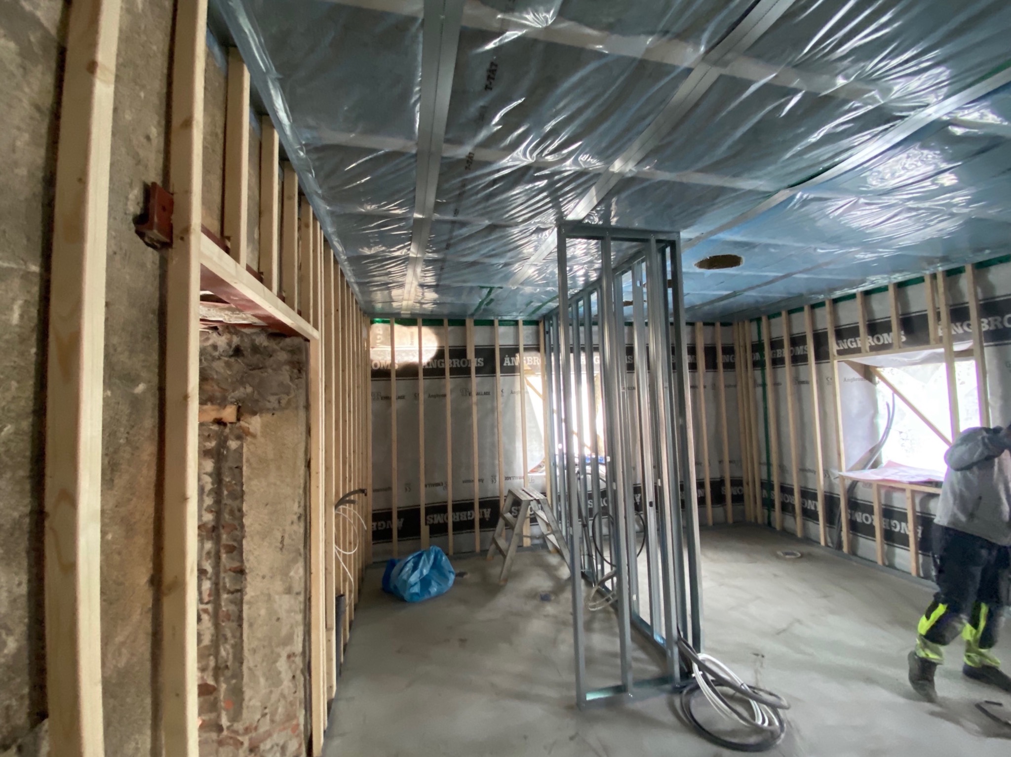 Wnętrze w trakcie prac wykończeniowych: drewniany szkielet ścian, folia na suficie, metalowa konstrukcja ścianki działowej, widoczne przewody i drabina. Fragment ściany z cegły i instalacją...