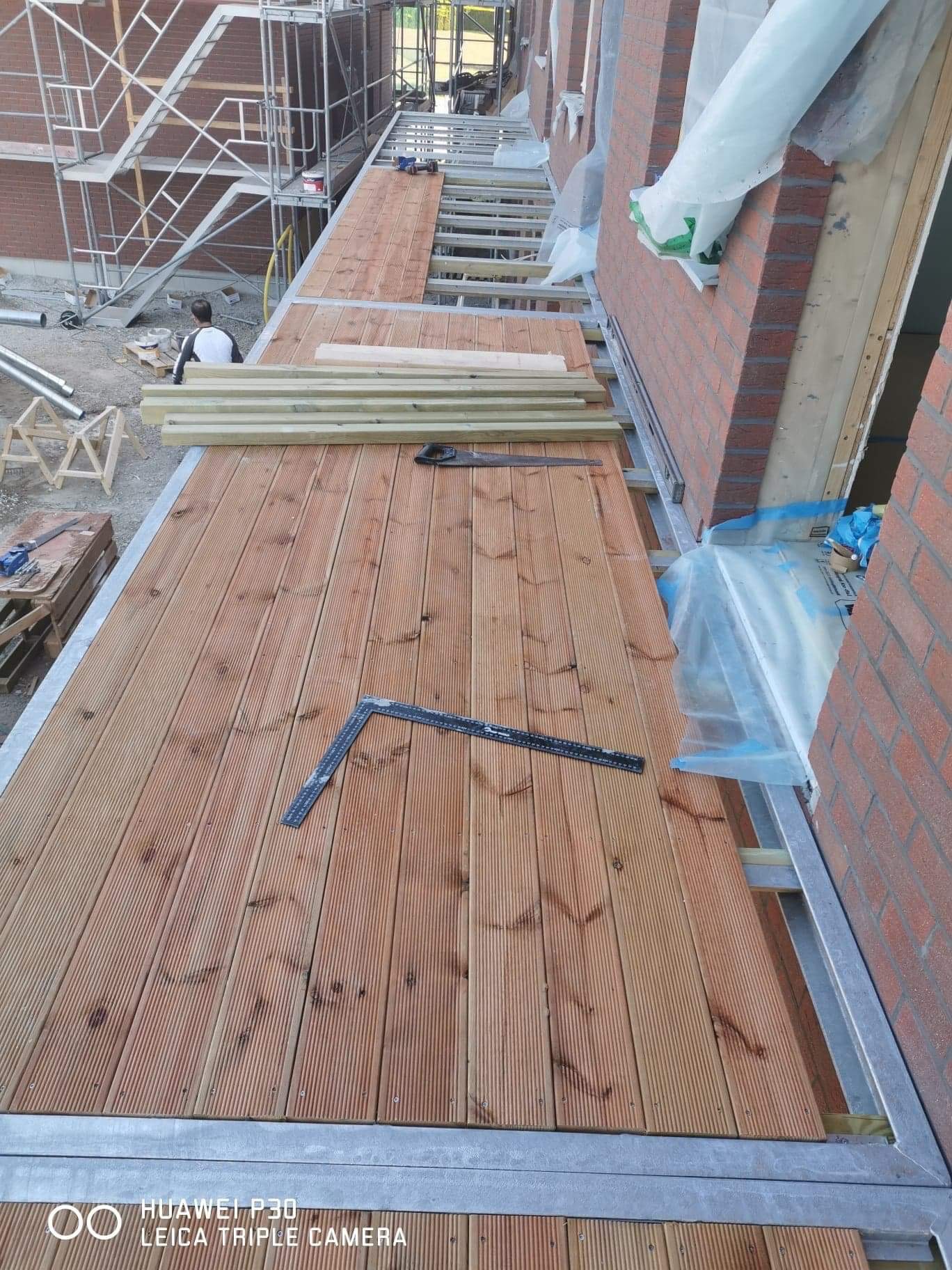 Montaż drewnianego tarasu na balkonie z widocznymi deskami, kątownikiem, piłą ręczną i rusztowaniami w tle, w trakcie prac wykończeniowych.