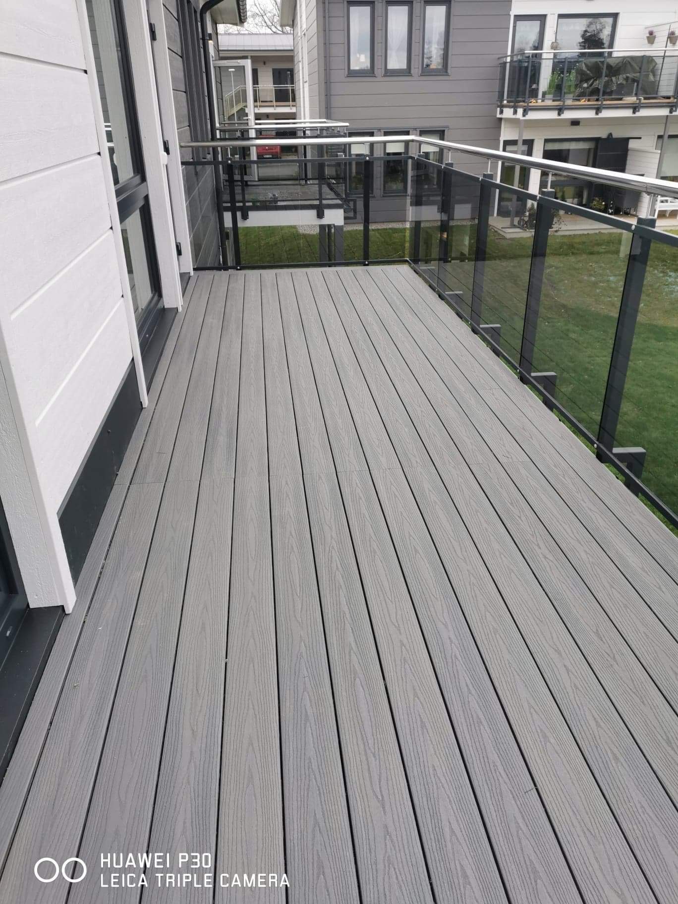 Nowoczesny balkon z szarą kompozytową podłogą z desek i szklaną balustradą z metalowymi ramami.