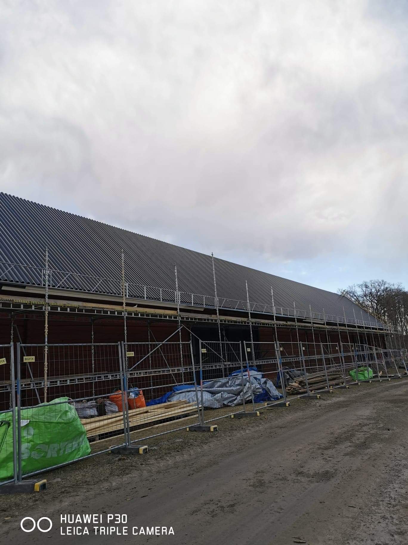 Budynek z nowym dachem z blachy trapezowej, zabezpieczony rusztowaniami i ogrodzeniem tymczasowym, widoczne materiały budowlane i narzędzia, pochmurne niebo.