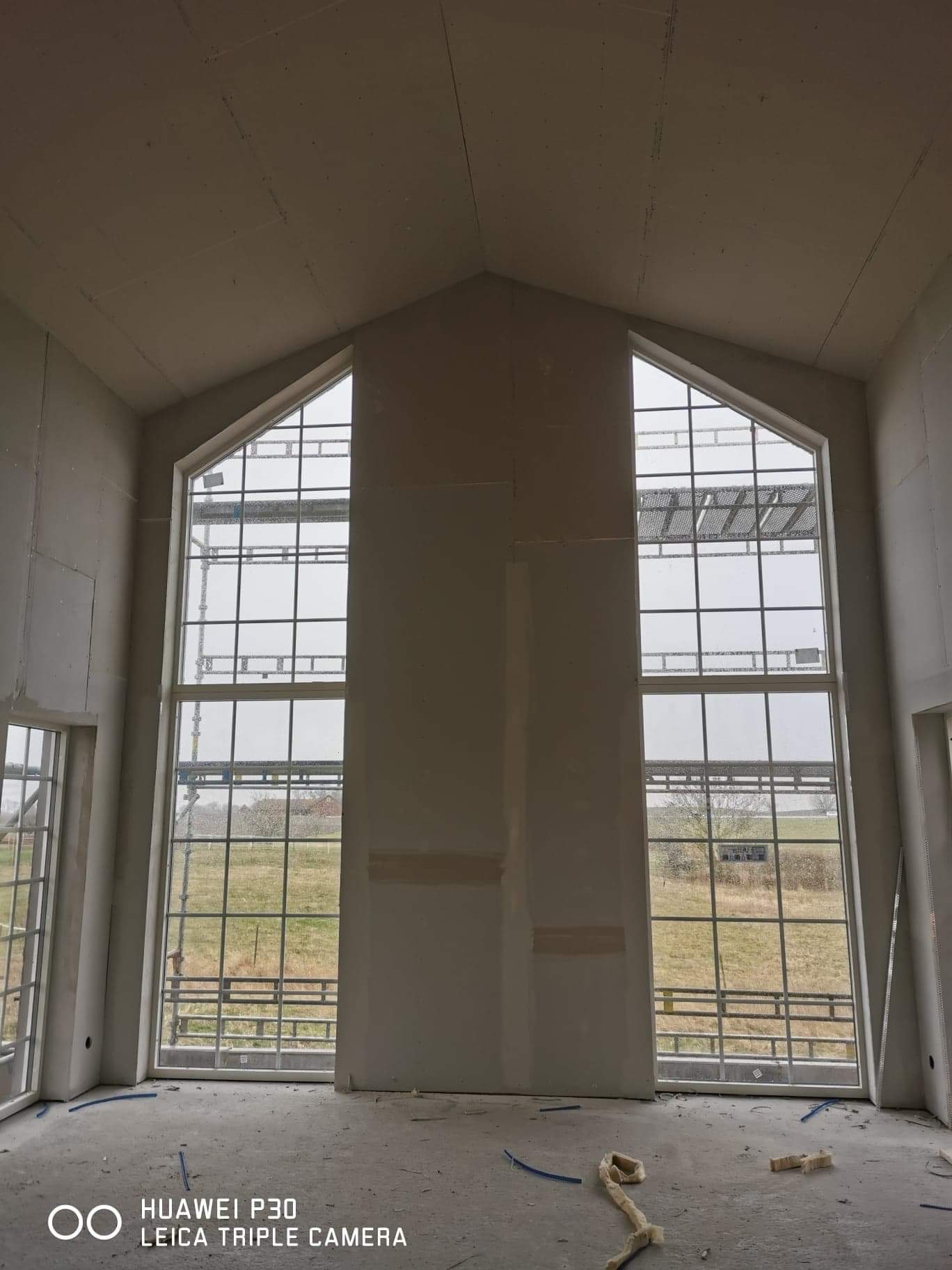 Wysokie okna z wieloma szprosami w surowym wnętrzu z płyt gipsowo-kartonowych, widoczne rusztowania na zewnątrz.