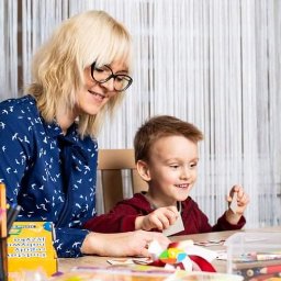 Neurologopeda - Kobieta w okularach i koszuli w ptaszki uśmiecha się do chłopca podczas ćwiczeń z kartami obrazkowymi na drewnianym stole, w tle firanka z cienkich nitek i pojemnik z pisakami.
