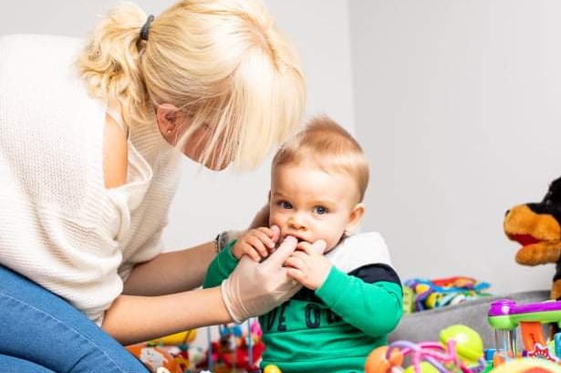Kobieta w rękawiczkach delikatnie sprawdza jamę ustną małego dziecka siedzącego wśród zabawek.