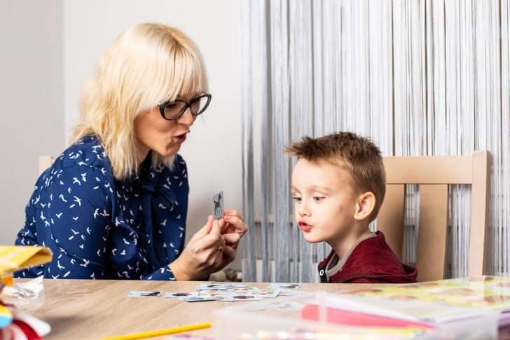 Kobieta w okularach i wzorzystej bluzce pokazuje dziecku kartę z obrazkiem konia podczas zajęć terapeutycznych przy drewnianym stole.