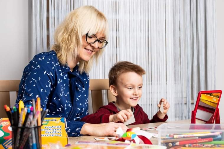 Kobieta w okularach i koszuli w ptaszki uśmiecha się do chłopca podczas ćwiczeń z kartami obrazkowymi na drewnianym stole, w tle firanka z cienkich nitek i pojemnik z pisakami.