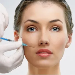 Kobieta podczas zabiegu iniekcji w policzek, widoczne ręce w białych rękawiczkach trzymające strzykawkę z niebieskim płynem, jasne tło.