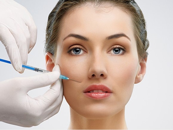 Kobieta podczas zabiegu iniekcji w policzek, widoczne ręce w białych rękawiczkach trzymające strzykawkę z niebieskim płynem, jasne tło.