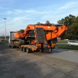 Pomarańczowa koparka gąsienicowa transportowana na niskopodwoziowej naczepie ciężarówki, widok z tyłu na tle błękitnego nieba.