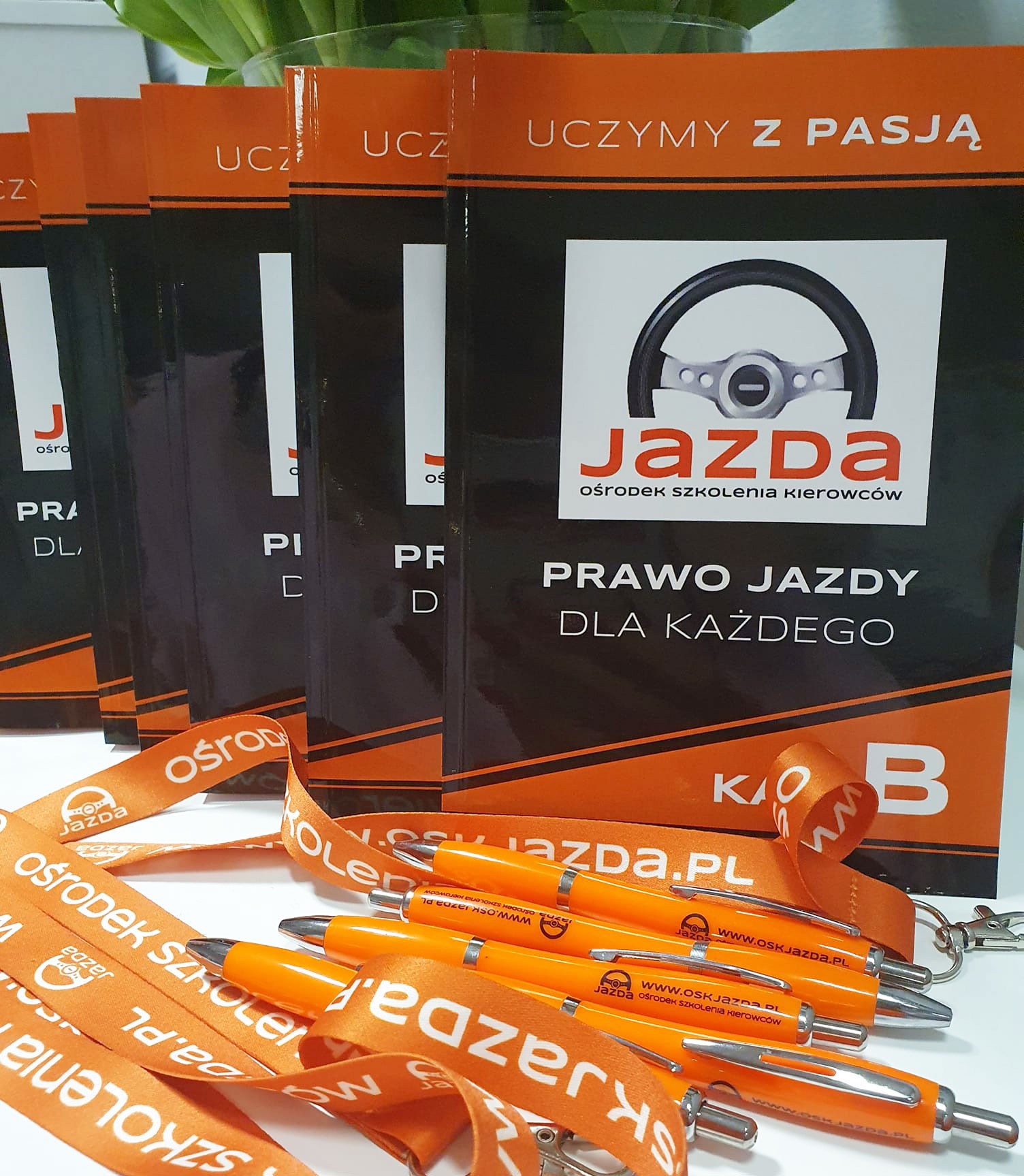 Promocyjne gadżety: pomarańczowe smycze i długopisy z logo ośrodka szkolenia kierowców, ułożone na tle książek 'Prawo jazdy dla każdego' z grafiką kierownicy.
