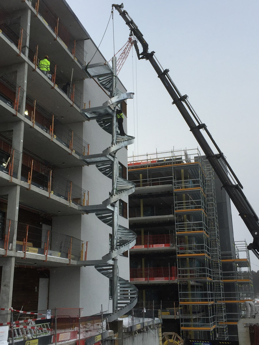 Montaż zewnętrznych schodów spiralnych na elewacji budynku w trakcie budowy za pomocą dźwigu, pracownik w odblaskowej kamizelce na schodach.