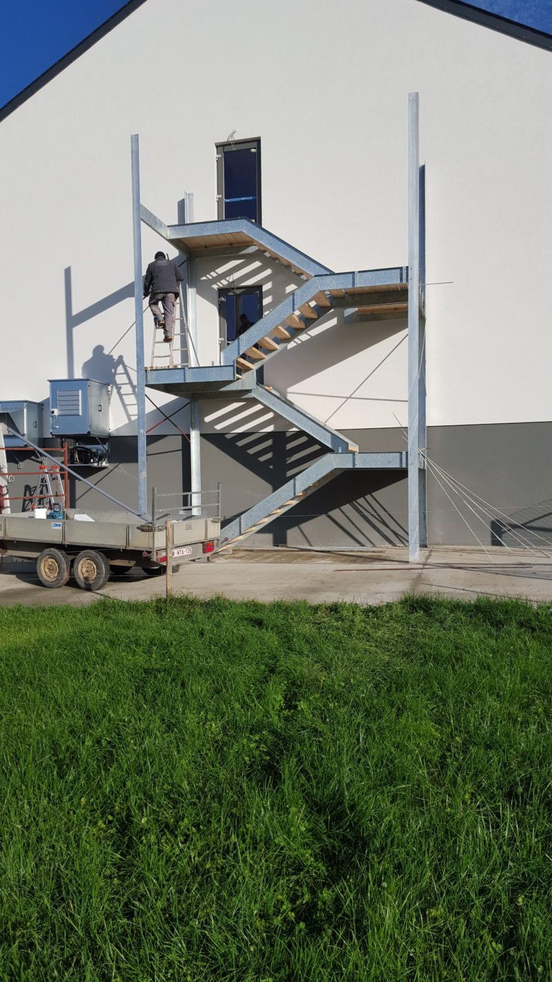 Zewnętrzna konstrukcja schodów z metalu przy białym budynku, pracownik na drabinie podczas montażu, widoczna przyczepa z narzędziami na trawniku.