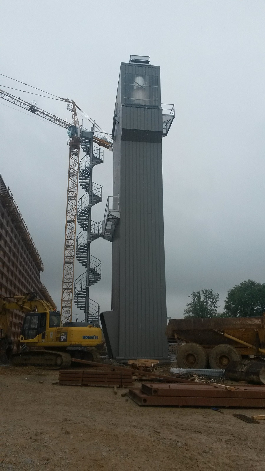 Metalowa wieża z zewnętrznymi schodami spiralnymi na placu budowy, widoczny dźwig i maszyny budowlane.