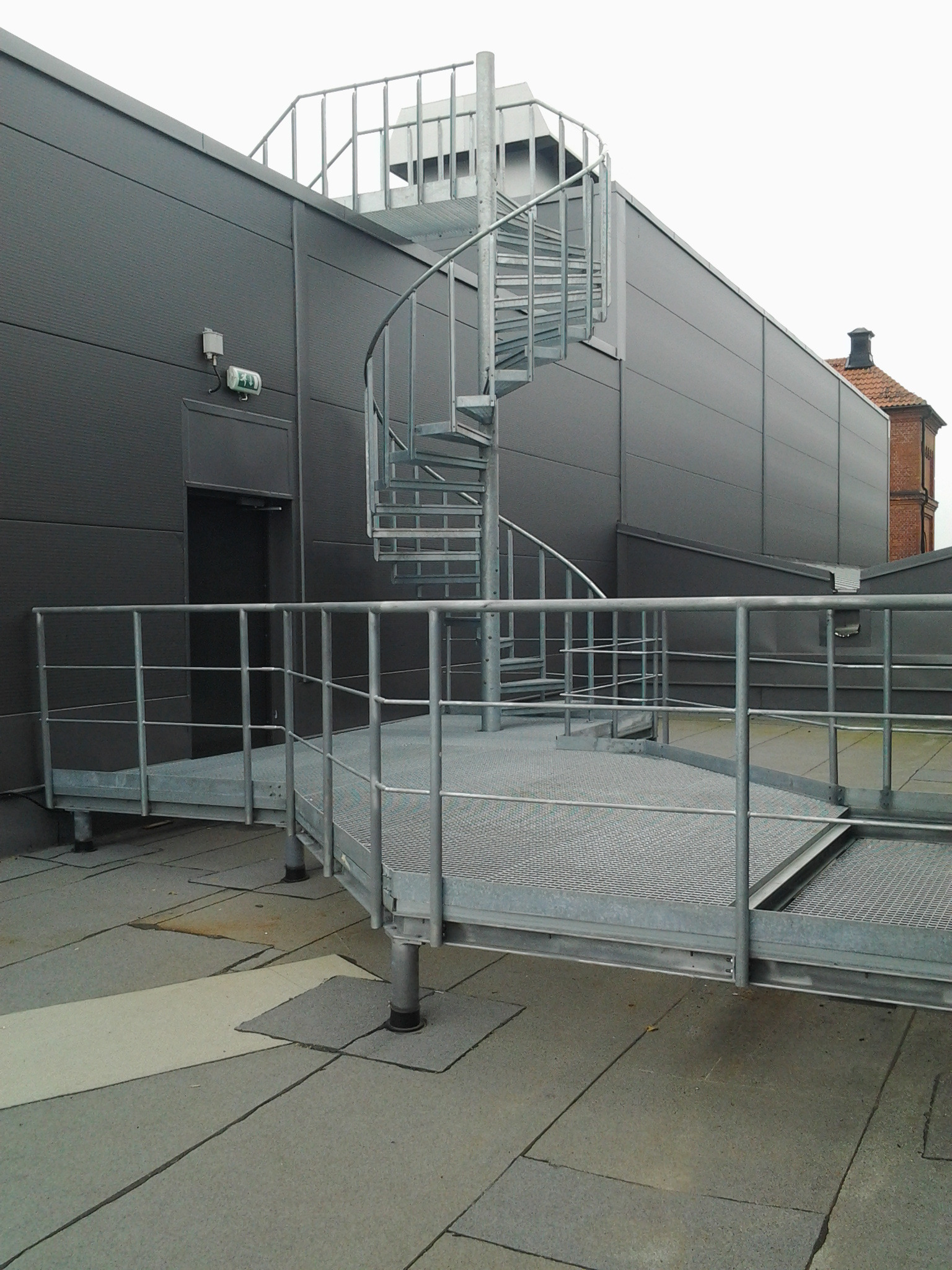 Zewnętrzne, spiralne schody ewakuacyjne z metalową platformą, prowadzące na dach budynku o szarej elewacji panelowej. Widoczny fragment budynku z czerwonej cegły w tle.