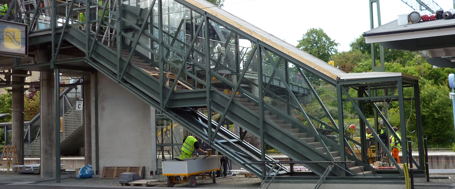 Zewnętrzna kładka dla pieszych z konstrukcji stalowej, częściowo oszklona, w trakcie montażu; widoczni pracownicy w odblaskowych kamizelkach, narzędzia i materiały budowlane.