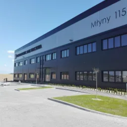 Nowoczesny budynek biurowo-magazynowy z ciemną elewacją, numerem 'Młyny 115' i nazwą 'IMPERIAL LOGISTIC PARK KORCZOWA', otoczony kostką brukową i małymi trawnikami z młodymi drzewkami.