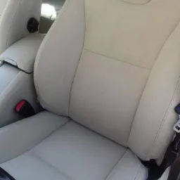 Jasny, beżowy fotel samochodowy z widocznym pasem bezpieczeństwa i fragmentem kierownicy.