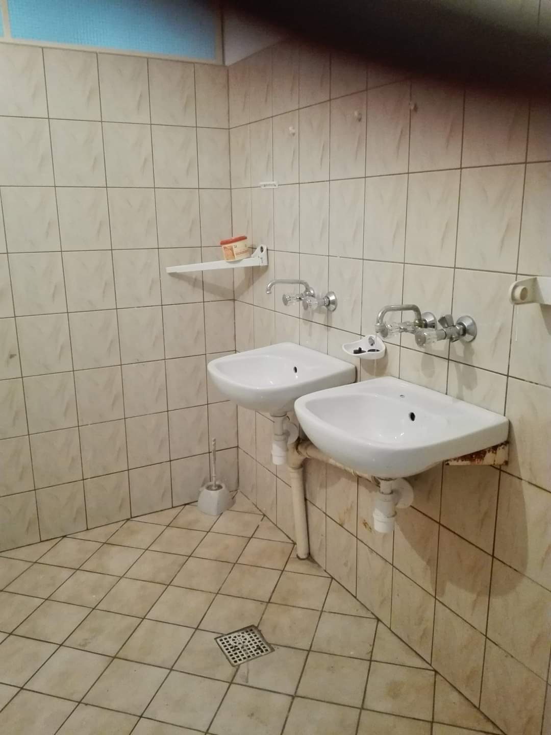 Dwie białe umywalki zamontowane na ścianie wyłożonej kwadratowymi, beżowymi płytkami. Widoczny odpływ podłogowy i szczotka do WC. Umywalki z widocznymi śladami użytkowania.