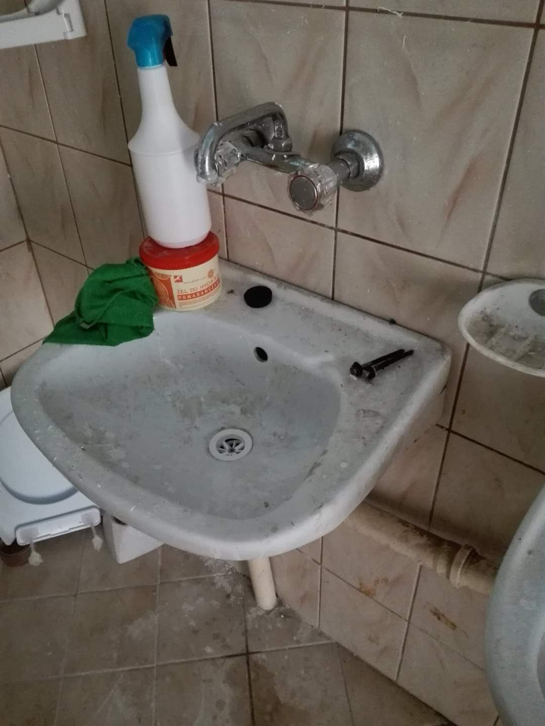 Brudna, narożna umywalka w łazience z widocznymi śladami użytkowania, butelka ze spryskiwaczem, słoik z żelem do mycia, zielona ściereczka i narzędzia leżące na brzegu umywalki.