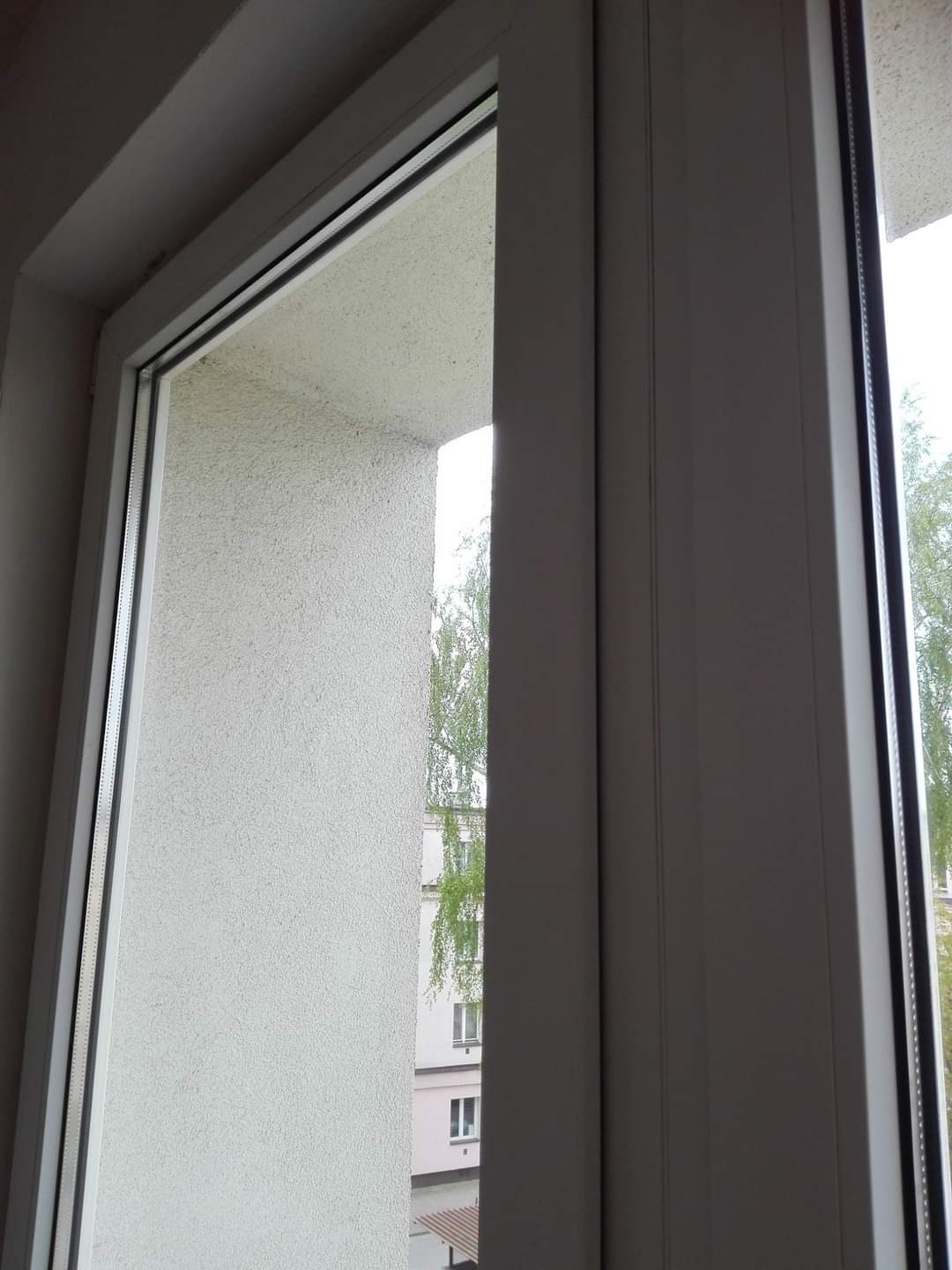 Widok przez otwarte białe okno na budynek z zielenią, widoczna faktura ściany i rama okienna.