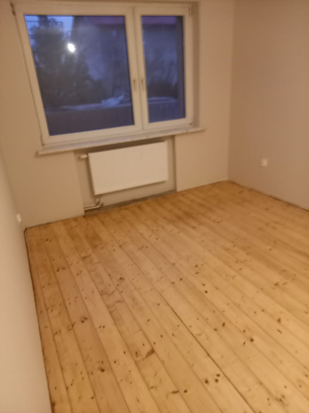 Pusty pokój z drewnianą podłogą, grzejnikiem pod oknem i ścianami w kolorze beżowym, przygotowany do dalszych prac wykończeniowych.