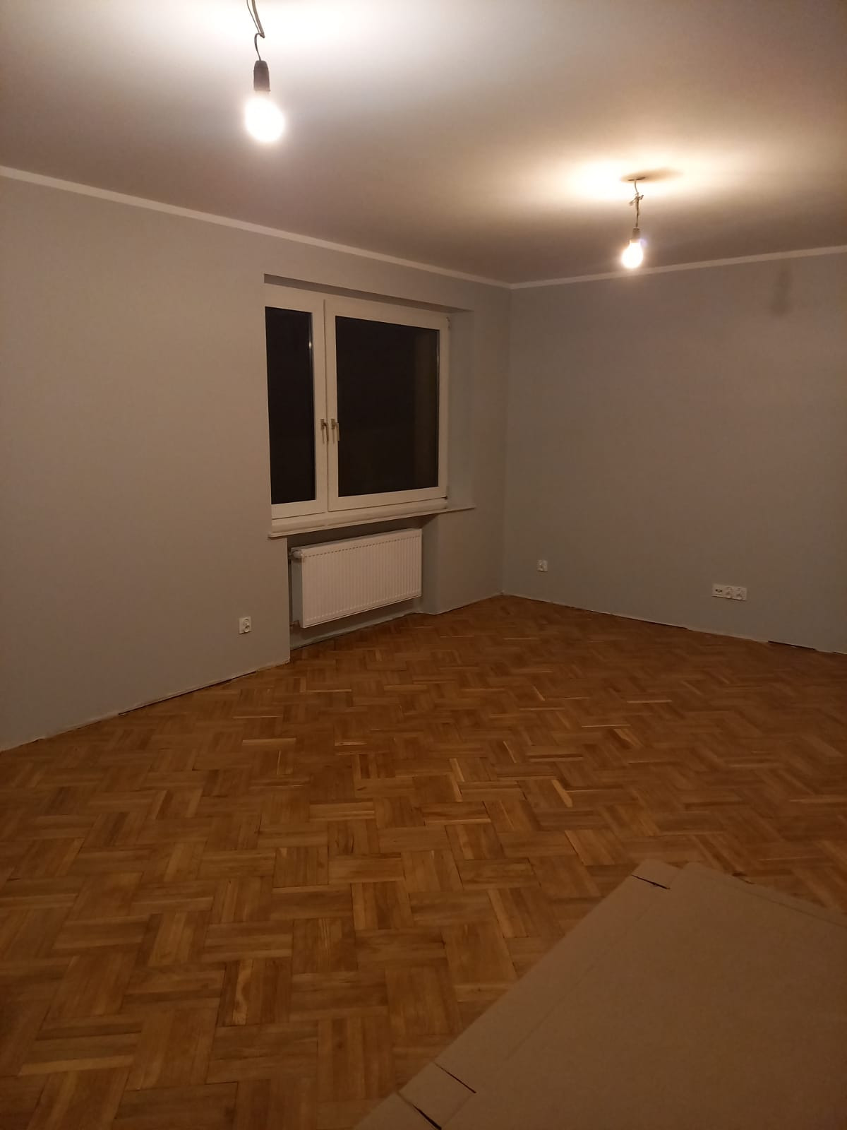 Szare ściany w pokoju z parkietem w jodełkę, okno z białymi ramami i wiszące żarówki, po świeżym malowaniu.