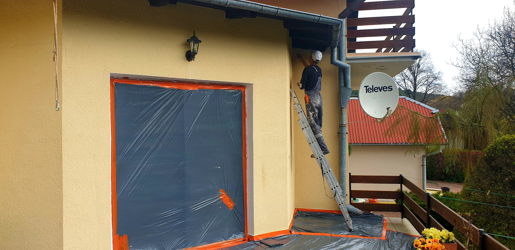 Pracownik na drabinie maluje elewację domu, okno zabezpieczone folią, widoczna antena satelitarna.