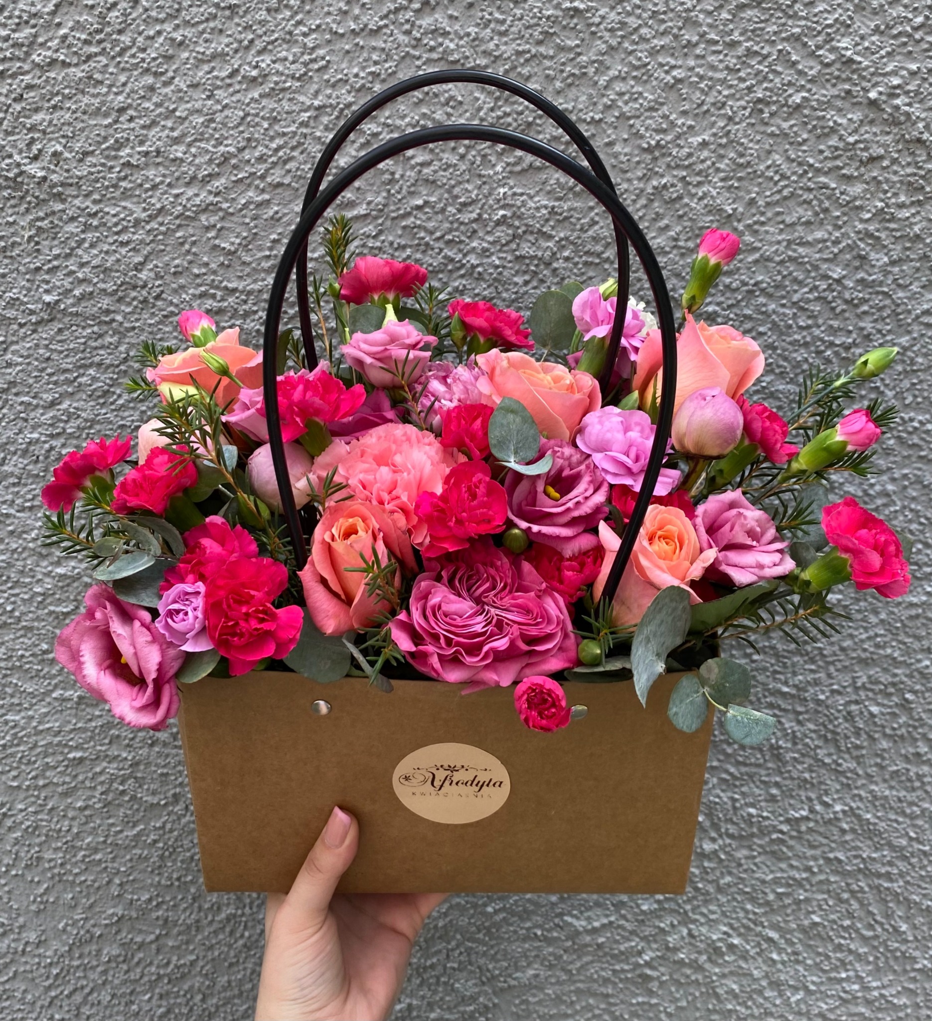 Ręcznie trzymany kosz prezentowy z różowymi i pomarańczowymi różami, goździkami i zielonymi dodatkami na tle szarej ściany z fakturą, z naklejką kwiaciarni.
